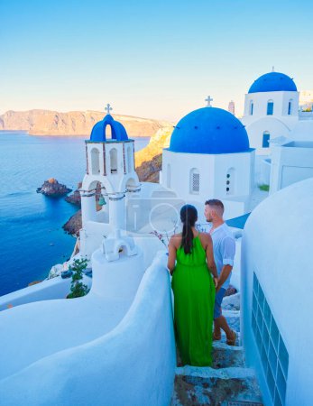 Foto de Pareja de vacaciones en Santorini Grecia, hombres y mujeres en el pueblo griego de Oia con una vista sobre el océano durante las vacaciones de verano viendo la puesta de sol una iglesia cúpula azul en las calles estrechas - Imagen libre de derechos