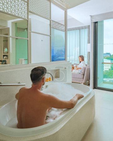 Foto de Una habitación de hotel con colores frescos brillantes, un dormitorio de estilo minimalista con vista al mar, hombres y una bañera y una mujer tomando café mirando por la ventana al océano y la playa - Imagen libre de derechos