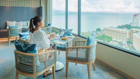 Photo for Asian Thai woman eating breakfast in a luxury hotel in Thailand, women drinking coffee looking out the window over the city and ocean - Royalty Free Image
