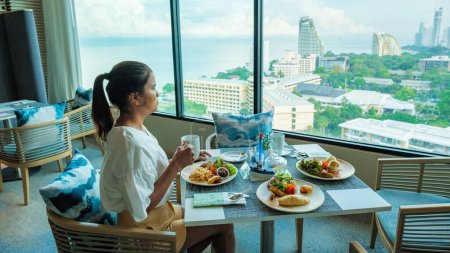 Photo for Asian Thai woman eating breakfast in a luxury hotel in Thailand, women drinking coffee looking out the window over the city of Pattaya and ocean - Royalty Free Image