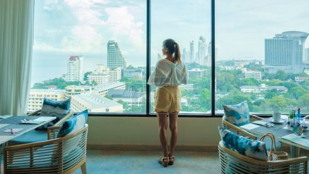 Photo for Asian Thai woman drinking coffee in the morning looking out the window of a skyscraper during breakfast in a luxury hotel in Pattaya Thailand - Royalty Free Image