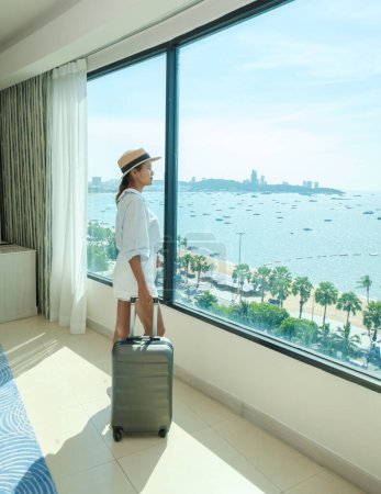Foto de Una habitación de hotel con colores frescos brillantes, un dormitorio de estilo minimalista con vista al mar, una mujer con equipaje de mano registrándose en una habitación de hotel, una mujer asiática con maleta - Imagen libre de derechos