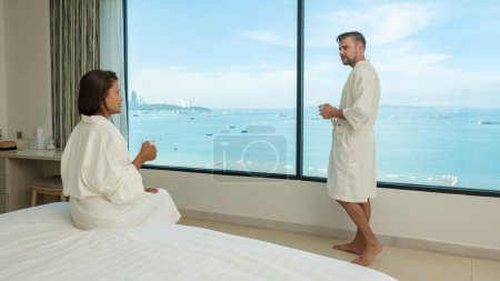 Téléchargez les photos : Un couple en vacances boire du café le matin en regardant par la fenêtre sur la plage et l'océan pendant des vacances de luxe - en image libre de droit