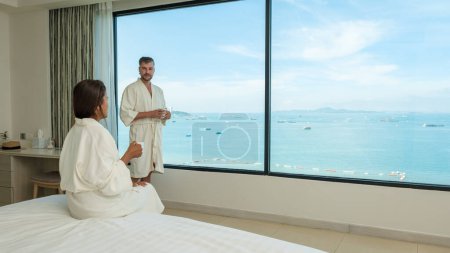 Téléchargez les photos : Couple en peignoir de bain en vacances boire du café le matin en regardant par la fenêtre sur la plage et l'océan pendant des vacances de luxe - en image libre de droit