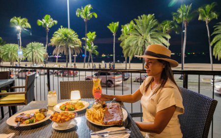Foto de Pattaya Tailandia 10 noviembre 2023, Hard rock café y hotel en la carretera de la playa al atardecer con cócteles en la mesa, mujer cenando por la noche - Imagen libre de derechos