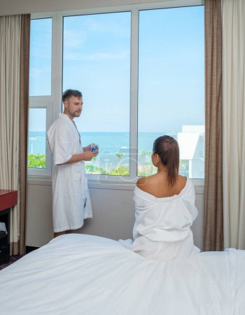 Téléchargez les photos : Un couple en vacances boire du café le matin regardant par la fenêtre sur la plage et l'océan pendant des vacances de luxe en Thaïlande - en image libre de droit