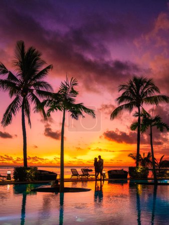 Téléchargez les photos : Un couple d'hommes et de femmes regardant le coucher du soleil sur une plage tropicale à Maurice avec des palmiers près de la piscine, le coucher du soleil tropical sur la plage à Maurice, un couple en voyage de lune de miel - en image libre de droit