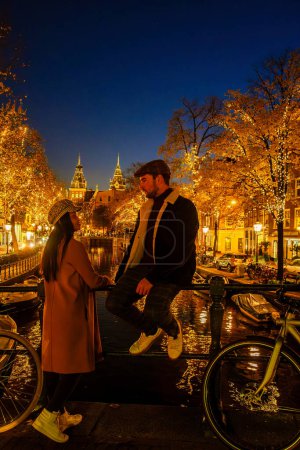 Téléchargez les photos : Amsterdam Pays-Bas canaux avec lumières de Noël en Décembre, centre historique du canal d'Amsterdam la nuit.Un couple d'hommes et une femme sont en voyage en ville pendant l'hiver - en image libre de droit