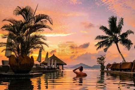 Téléchargez les photos : Piscine de luxe dans une station balnéaire tropicale, vacances relaxantes dans les îles Seychelles. La Digue, Jeune homme au coucher du soleil au bord de la piscine Seychelles, hommes regardant le coucher du soleil depuis la piscine avec des palmiers - en image libre de droit