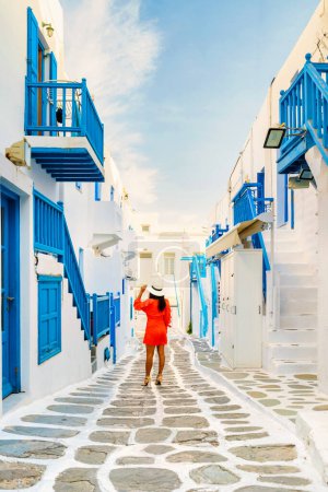 Foto de Mujeres jóvenes en vestido rojo caminan por la calle del pueblo griego de Mykonos en Grecia, calles coloridas del pueblo de Mikonos. Mujer asiática visita Mykonos pueblo durante vacaciones en Europa - Imagen libre de derechos
