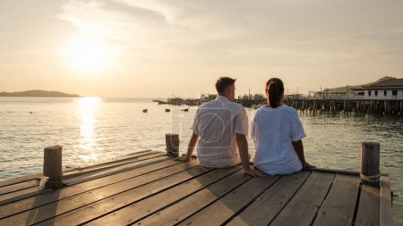 Téléchargez les photos : Un couple d'hommes européens et une femme asiatique en vacances en Thaïlande, couple assis sur une jetée de pont en bois dans l'océan pendant le coucher du soleil en Thaïlande samésienne - en image libre de droit