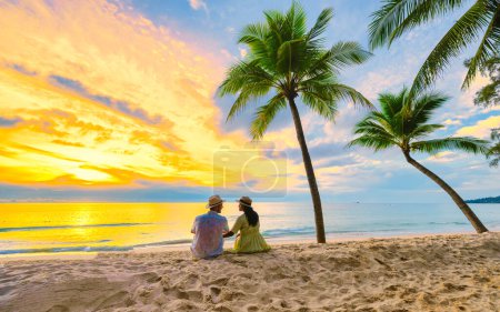 Foto de Un par de hombres y mujeres sentados en la playa viendo la puesta de sol en la playa con arena blanca y palmeras, Bang Tao playa Phuket Tailandia. - Imagen libre de derechos