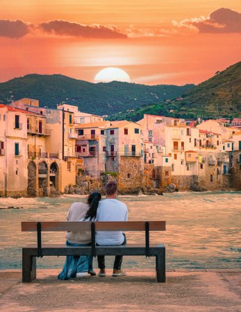 Photo for A couple of men and woman sitting on a bench at the waterfront on vacation in Sicily during sunset, a couple visiting the old town of Cefalu Sicilia Italy - Royalty Free Image