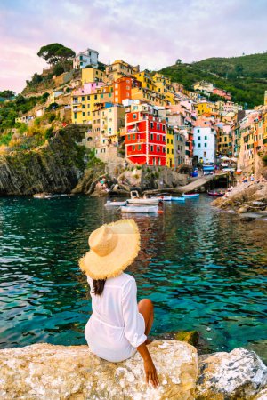 Téléchargez les photos : Riomaggiore Cinque Terre Italie, port de village coloré face à l'océan, jeune femme asiatique avec un chapeau regardant le coucher du soleil au bord de l'eau donnant sur le village de Riomaggiore Cinque Terre Italie - en image libre de droit