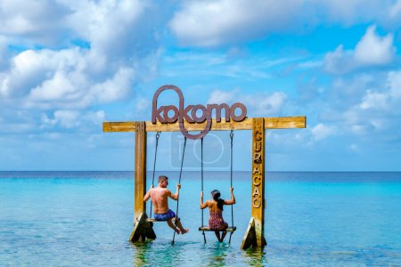 Téléchargez les photos : Couple d'hommes et de femmes à la balançoire dans l'océan de l'île des Caraïbes de Curaçao, Kokomo Beach à l'île des Caraïbes de Curaçao par une journée ensoleillée - en image libre de droit