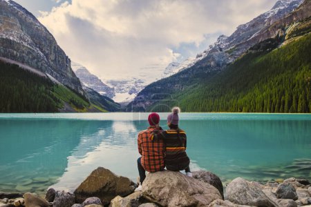 Téléchargez les photos : Lake Louise Banff National Park in the Canadian Rocky Mountains. Un jeune couple d'hommes et de femmes assis sur un rocher au bord du lac par une journée froide à l'automne au Canada regardant le coucher du soleil - en image libre de droit