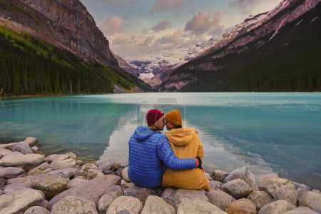 Téléchargez les photos : Lake Louise Banff National Park in the Canadian Rocky Mountains. Un jeune couple d'hommes et de femmes assis sur un rocher au bord du lac par une journée froide à l'automne au Canada regardant le coucher du soleil au lac - en image libre de droit