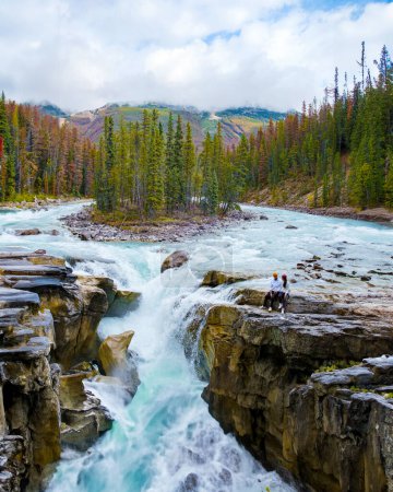 Foto de Sunwapta Falls Jasper National Park Canada. The Canadian Rockies during the Autumn fall season. Un par de hombres y mujeres visitando Sunwapta Falls de pie en el borde de las rocas junto al río - Imagen libre de derechos