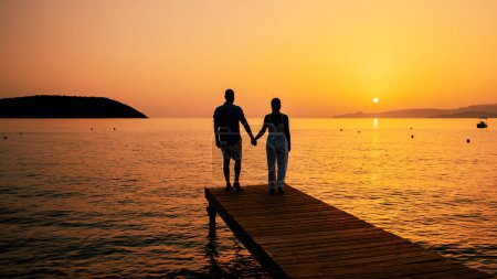 Téléchargez les photos : Un couple d'hommes et de femmes sur une jetée en bois regardant le coucher de soleil sur la mer, hommes et femmes regardant un coucher de soleil en Crète Grèce Europe sur une jetée en bois dans l'océan - en image libre de droit