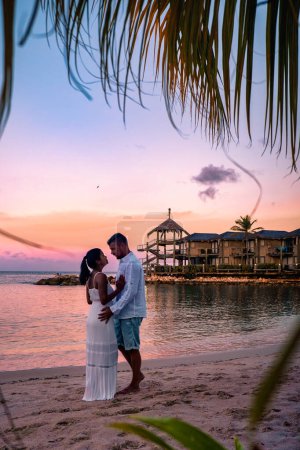 Téléchargez les photos : Resort de luxe tropical Curaçao avec plage privée et palmiers, vacances de luxe Curaçao Caraïbes, couple hommes et femmes regardant coucher de soleil à la plage avec piscine et palmiers - en image libre de droit