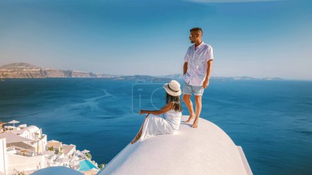 Foto de Santorini Grecia, una joven pareja de vacaciones de lujo en la isla Santorini observando el amanecer por la iglesia de cúpula azul y el pueblo encalado Oia Santorini, mujeres asiáticas y hombres caucásicos de vacaciones - Imagen libre de derechos