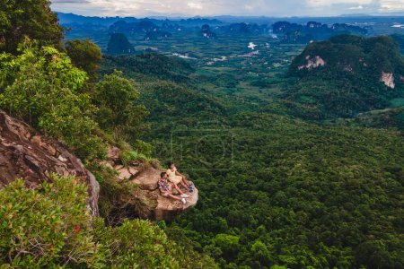 Téléchargez les photos : Dragon Crest Mountain Krabi Thaïlande, un jeune couple est assis sur un rocher qui surplombe l'abîme avec un beau paysage. Dragon Crest ou Khuan Sai au Khao Ngon Nak Nature Trail dans la forêt tropicale de Krabi - en image libre de droit