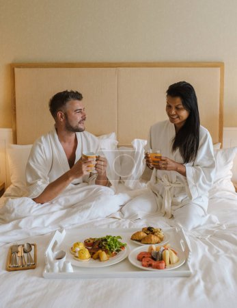 Photo for Couple having breakfast in bed at a luxury hotel, men and women in bed with breakfast coffee, and bread. a diverse couple of an Asian woman and a European man in bedroom - Royalty Free Image