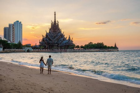 Téléchargez les photos : Un couple multiethnique diversifié d'hommes et de femmes visitent le temple en bois du Sanctuaire de la Vérité à Pattaya en Thaïlande au coucher du soleil sur la plage - en image libre de droit
