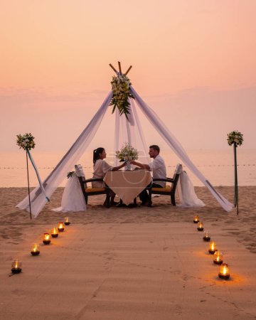 Téléchargez les photos : Dîner romantique sur la plage à Pattaya en Thaïlande, un couple homme et femme d'âge moyen asiatique, et un homme européen dînant sur la plage en Thaïlande pendant le coucher du soleil. - en image libre de droit