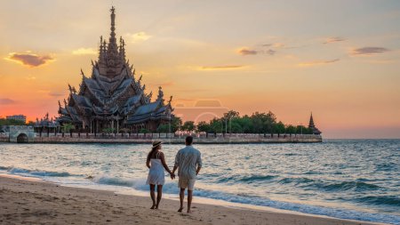 Téléchargez les photos : Un couple multiethnique diversifié d'hommes et de femmes visitent le temple en bois du Sanctuaire de la Vérité à Pattaya Thaïlande au coucher du soleil, couple regardant le coucher du soleil sur la plage à Pattaya avec un temple en bois - en image libre de droit