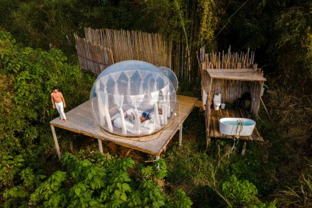 Téléchargez les photos : Couple homme et femmes dans une tente à bulles avec une salle de bain extérieure avec une baignoire dans la forêt tropicale de la jungle. Glamping de luxe - en image libre de droit