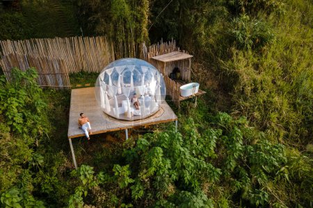 Téléchargez les photos : Couple homme et femmes dans une tente à bulles avec une salle de bain extérieure avec une baignoire dans la forêt tropicale de la jungle. Glamping de luxe en Thaïlande - en image libre de droit