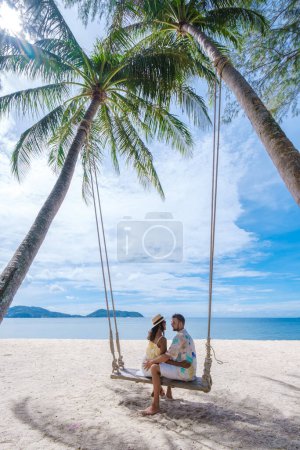 Téléchargez les photos : Un couple d'hommes et de femmes se détendent sur une plage tropicale blanche avec des palmiers à Phuket Thaïlande. hommes et femmes dans une corde balancent sous un palmier par une journée ensoleillée - en image libre de droit