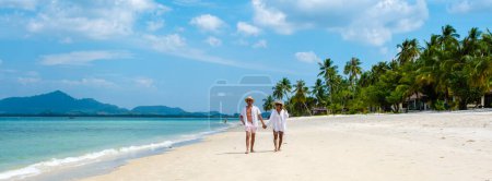 Photo for A couple of men and woman with summer hats walking at the beach of Koh Muk a tropical island with palm trees soft white sand, and a turqouse colored ocean in Koh Mook Trang Thailand - Royalty Free Image