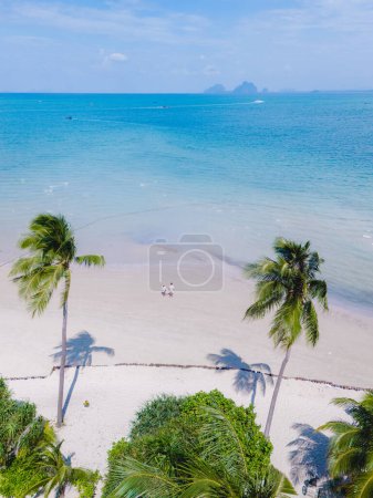 Téléchargez les photos : Vue entre les palmiers d'un couple marchant sur la plage tropicale de sable blanc de Koh Muk, palmiers sable blanc doux, et une turque couleur océan à Koh Mook Trang Thaïlande - en image libre de droit