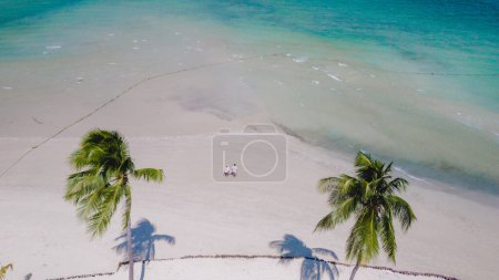 Photo for View between palm trees at a couple walking on the white sandy tropical beach of Koh Muk, palm trees soft white sand, and a turqouse colored ocean in Koh Mook Trang Thailand - Royalty Free Image