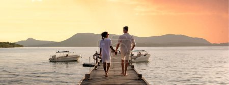 couple walking on a wooden pier in the ocean at sunset in Thailand.Caucasian men and Asian women diverse couple waking at a jetty in the ocean, man and women watching sunset together