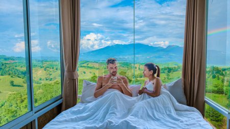 Photo for A couple of men and women laying in bed with open widows looking out over the mountains of Thailand, men and woman in bedroom with large open glass windows looking out over the green landscape of Nan - Royalty Free Image