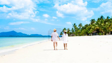 ein paar Männer und Frauen mit sommerlicher tropischer Insel Koh Muk, ein kaukasischer Mann und eine asiatische Frau, die im Sommerurlaub in Koh Mook Trang Thailand an einem weißen Strand mit Palmen spazieren gehen