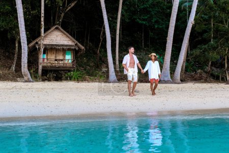 Téléchargez les photos : Couple diversifié de femme asiatique et d'homme européen marchant sur la plage au coucher du soleil, sur le fond un bungalow de cabane en bambou en bois. un jeune couple d'hommes et de femmes sur une île tropicale en Thaïlande - en image libre de droit