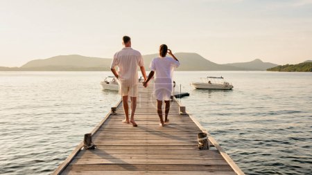 Photo for A man and a woman standing on a dock by a lake, holding hands. They are wearing shorts and looking out at the horizon, happy and relaxed during their leisurely travel in Thailand, a couple wooden pier - Royalty Free Image