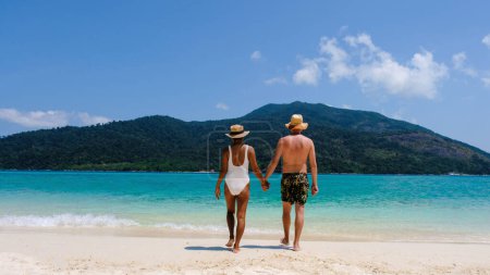 Téléchargez les photos : Vue arrière d'un couple sur la plage Koh Lipe Island Thaïlande, île tropicale avec un océan bleu et sable blanc doux. hommes et femmes se relaxant sur la plage marchant vers l'océan bleu, bikini et maillot de bain court - en image libre de droit