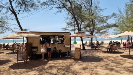 Téléchargez les photos : Rayong Thaïlande 13 mars 2024, Un groupe diversifié de personnes dégustant des aliments fraîchement préparés à partir d'un camion alimentaire lumineux sur une plage ensoleillée. - en image libre de droit