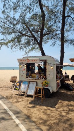 Foto de Rayong Tailandia 13 marzo 2024, Un vibrante camión de comida está estacionado en una playa de arena junto a un árbol exuberante, que sirve deliciosas golosinas para los asistentes a la playa bajo el cielo azul claro. - Imagen libre de derechos
