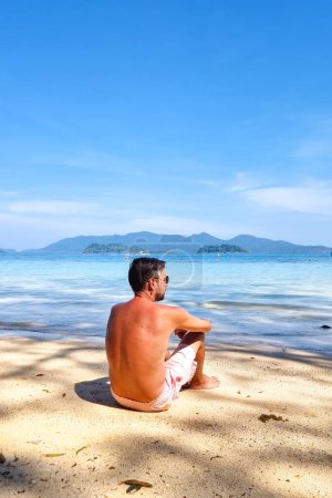 Téléchargez les photos : Un homme en tenue décontractée est assis sur la plage de sable, son regard fixé sur la vaste étendue de l'océan devant lui. Koh Wai Thaïlande - en image libre de droit