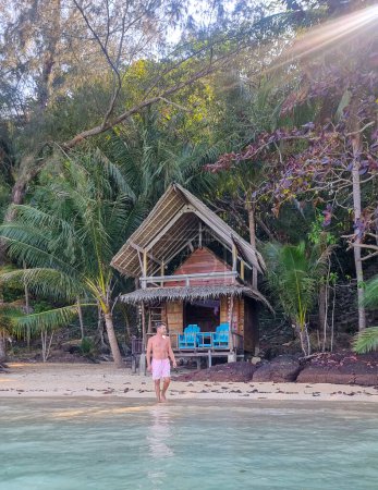 Téléchargez les photos : Un homme debout paisiblement sur une plage ensoleillée en face d'une cabane traditionnelle, trempant dans la beauté et la sérénité des environs. Koh Wai Thaïlande - en image libre de droit