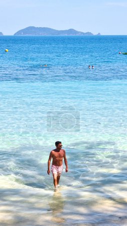 Téléchargez les photos : Un homme à l'apparence robuste sort gracieusement de l'eau scintillante de l'océan, les gouttelettes scintillent à la lumière du soleil alors qu'il émerge sur la plage de sable. Koh Wai Thaïlande - en image libre de droit