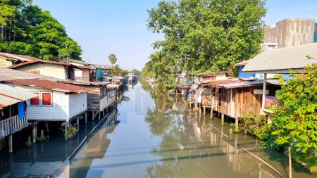 Téléchargez les photos : Bangkok Thaïlande 13 janvier 2024, Une rivière tranquille serpentant à travers une verdure luxuriante avec des maisons pittoresques perchées le long de ses rives, créant une scène pittoresque de bord de rivière vivant en harmonie avec la nature. - en image libre de droit