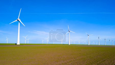 Foto de Una vasta extensión de tierra en Flevoland, Países Bajos, está cubierta con una plétora de molinos de viento imponentes girando al unísono bajo el vibrante cielo de primavera. turbinas de molino de viento energía verde - Imagen libre de derechos