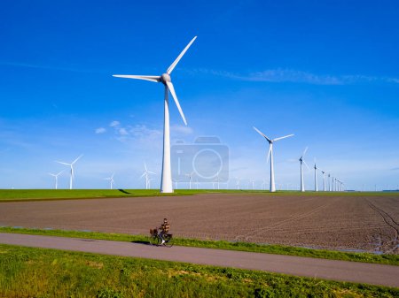 Téléchargez les photos : Un homme conduit son vélo électrique sur un chemin de terre le long d'éoliennes imposantes par une journée ensoleillée aux Pays-Bas Flevoland au printemps. hommes sur vélo électrique campagne - en image libre de droit